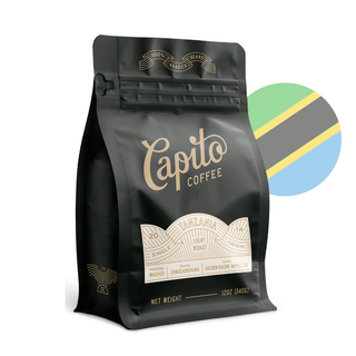 A black coffee bag labeled "Capito Coffee Origins Roast // Tanzania // Estate" with golden text. This 12 oz (340g) light roast from Tanzania features Arabica coffee with tasting notes of golden raisin, maple, and fig. Behind the bag is a circularly shaped flag of Tanzania, highlighting its unique flavor profile.