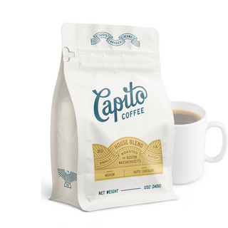  A 12 oz (340g) white bag of Capito Coffee House Blend sits next to a white mug filled with coffee. The bag features gold and blue accents with text indicating it is a medium roast with nutty and chocolate notes, roasted in small batches in Boston, Massachusetts.