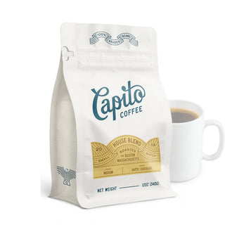 A white bag of Capito Coffee Case of House Blend stands beside a white mug of coffee, showcasing this classic Boston-roasted medium roast with nutty and chocolatey notes.